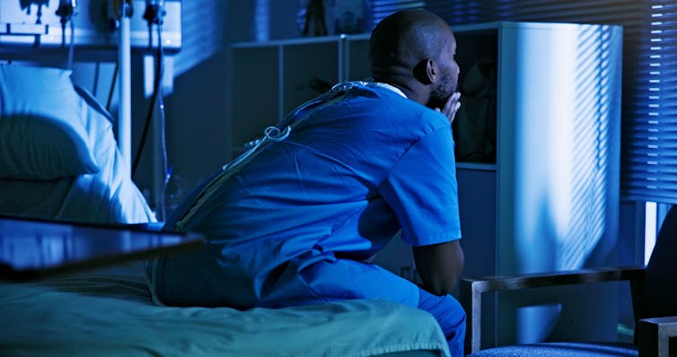 A patient in a gown sits on the edge of a hospital bed at night, with his elbows on his knees and his chin crossed