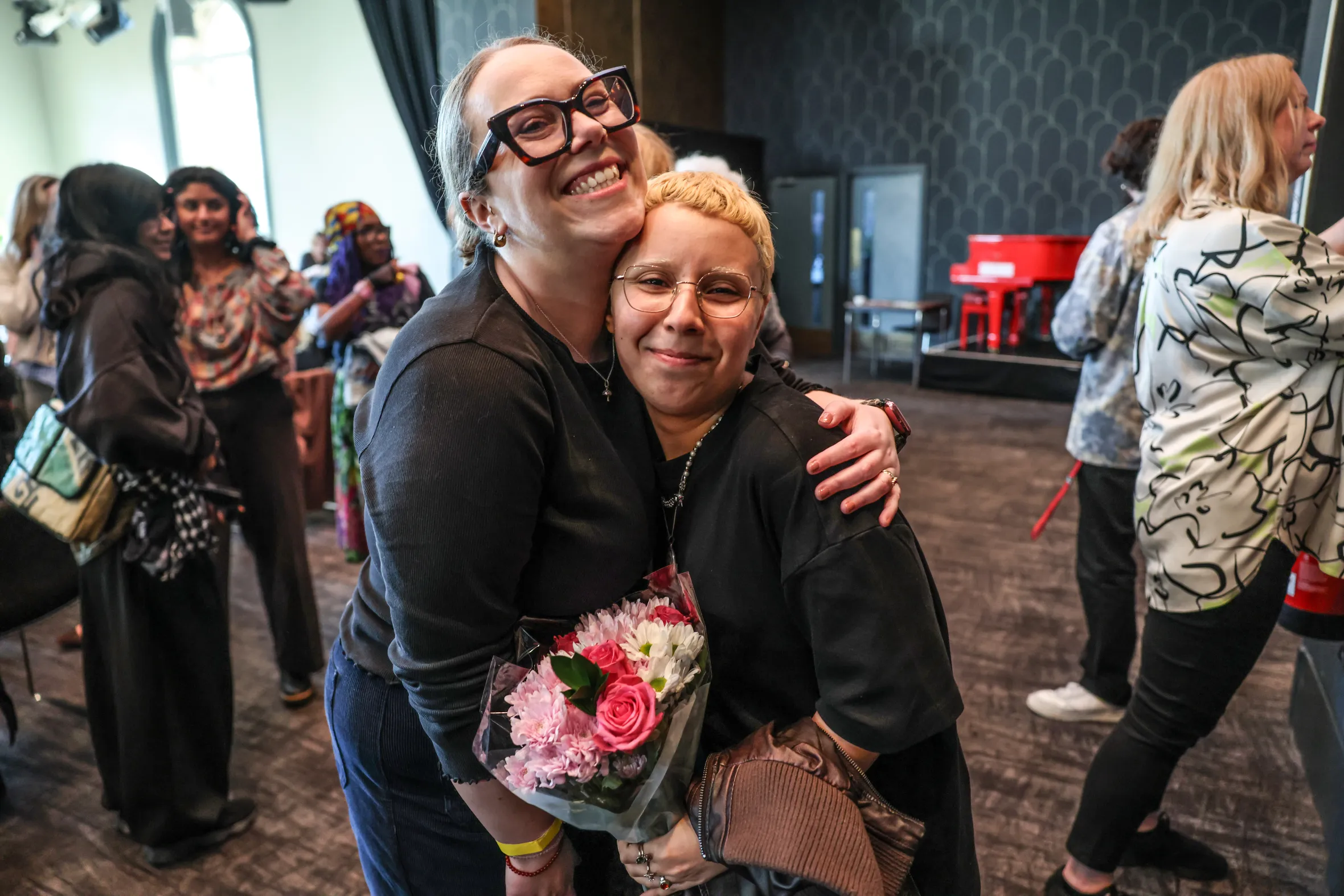 Carly Smallman hugs a woman with short blonde hair holding a bouquet of flowers after a stand-up comedy performance.