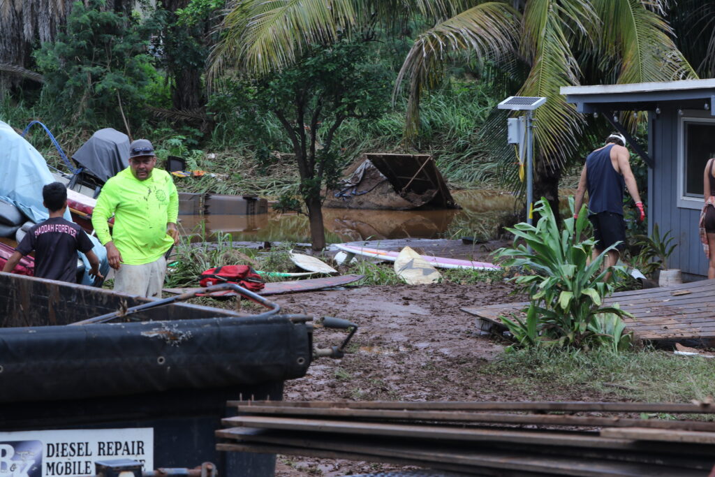 A man in a bright shirt stands next to a boy with a trash can and a board in the foreground. In the background is mud and a large pool of muddy water, with a garbage can floating in it.