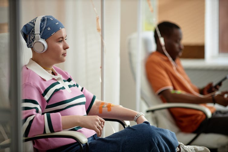 Two patients sitting in chairs with IVs attached to their arms, wearing casual clothes and headphones over their ears.