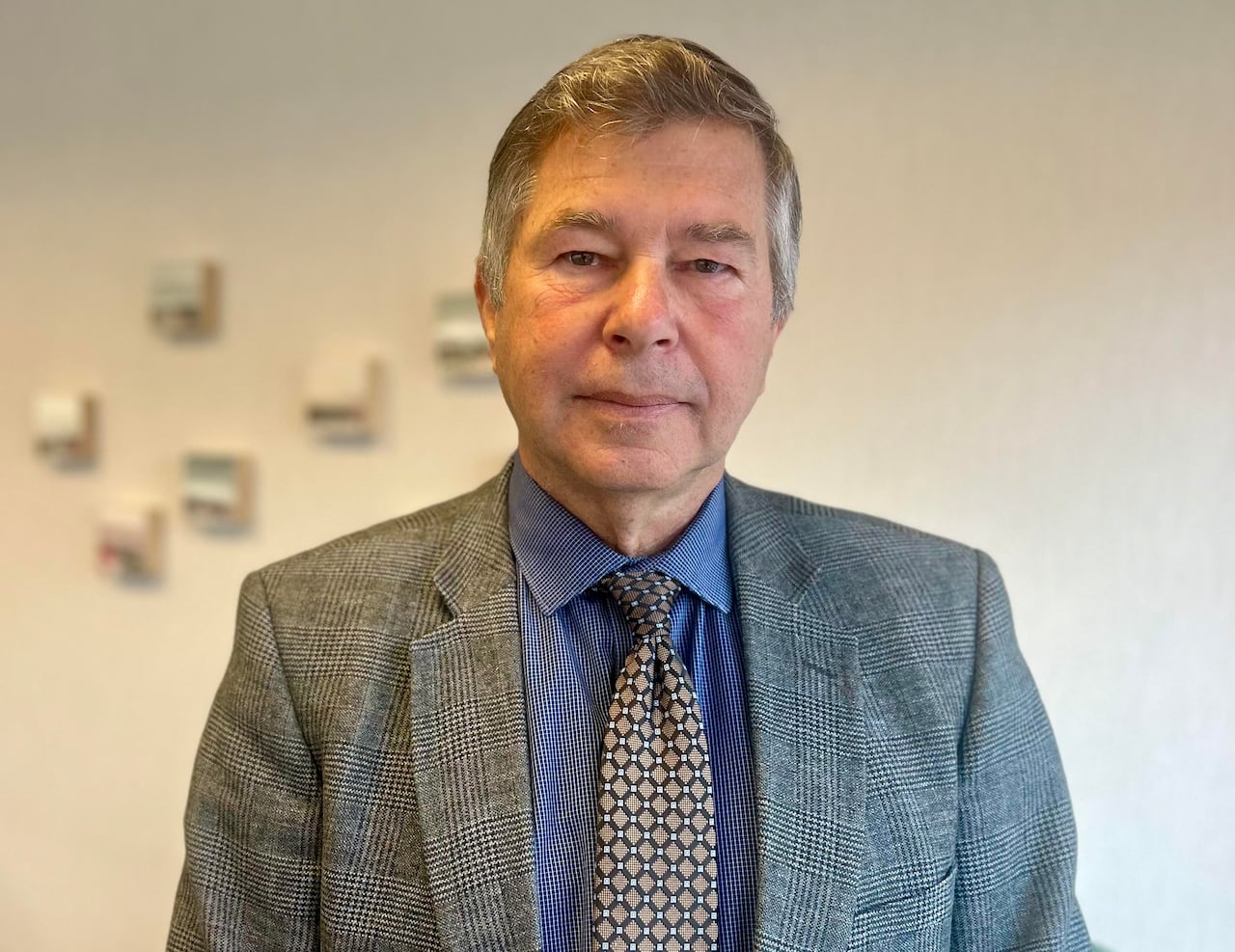 A white-haired man wearing a suit and tie stands in an office, staring at the camera with a serious face.  