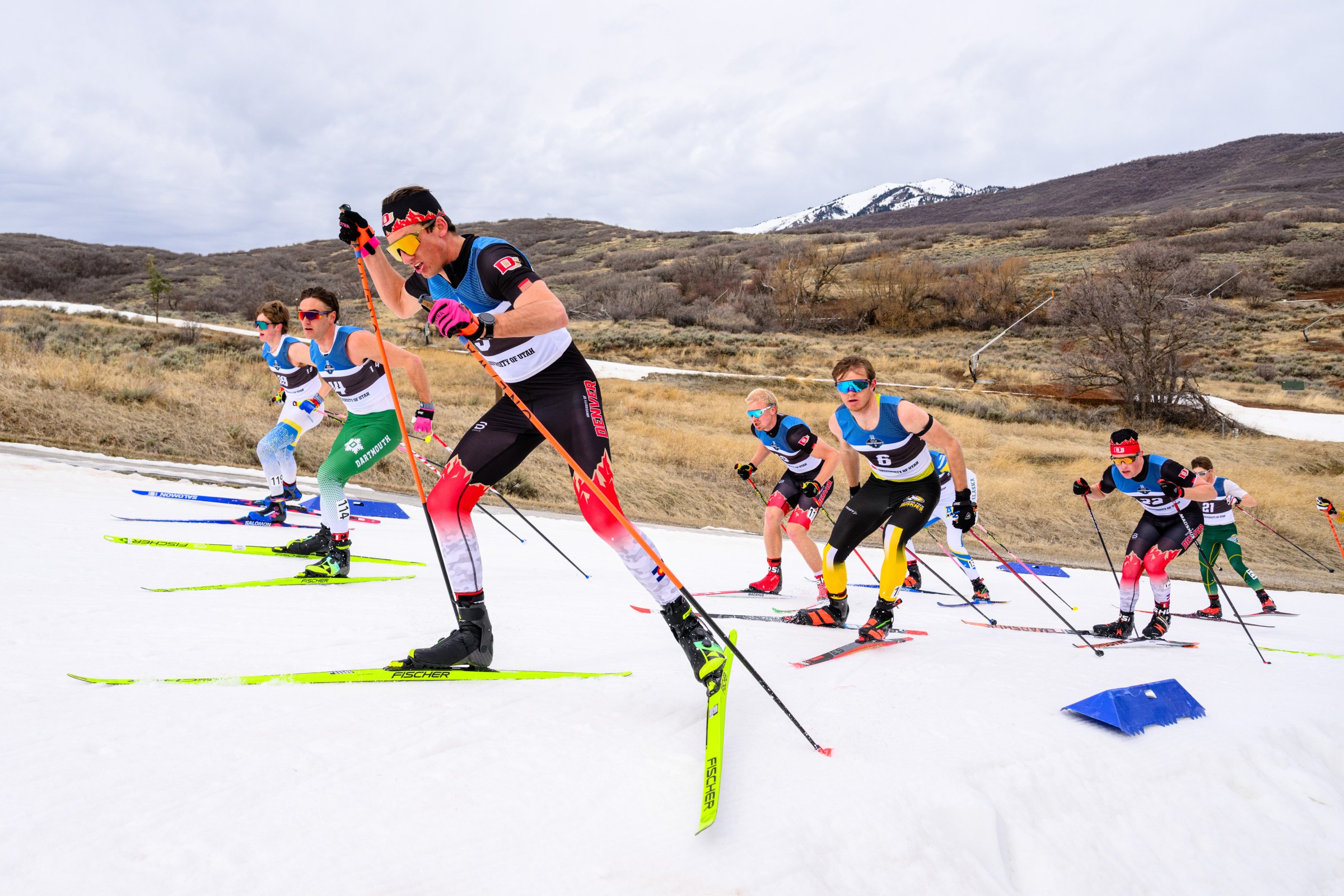 A college skier skis up a narrow snowy path with bare grassy hills around him