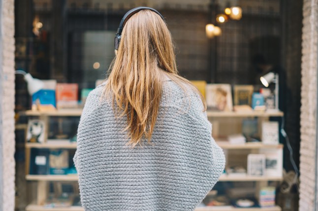 Young blond woman wearing headphones and a pullover on her back, looking out the window of a bookstore.